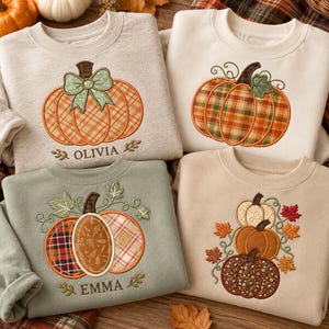 May include: Four crewneck sweatshirts in oatmeal, cream, and sage green colors, each featuring embroidered pumpkin designs. Two sweatshirts include personalized names, Olivia and Emma, stitched in dark brown thread below the pumpkins.