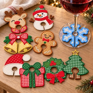 May include: A collection of felt Christmas wine glass charms featuring a reindeer, snowman, bells, gingerbread person, Santa hat, gift box, wreath, and tree. A blue snowflake charm is attached to the base of a wine glass filled with red wine.