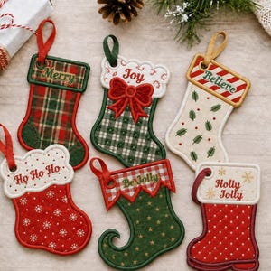 May include: Six embroidered Christmas stocking ornaments with hanging loops. Designs include a red and green plaid stocking labeled Merry, a green gingham stocking with a red bow labeled Joy, a cream stocking with pine needles labeled Believe, a red snowflake stocking labeled Ho Ho Ho, a green star stocking labeled Be Jolly, and a red quilted stocking labeled Holly Jolly.