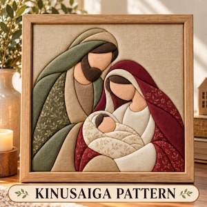 May include: A framed fabric wall art piece featuring a Kinusaiga pattern of a family. The figures are rendered in layered fabric textures with a sage green cloak, a deep red head covering, and a cream-colored swaddled infant in the center.