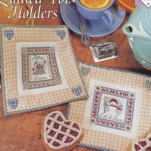 May include: A book cover featuring cross-stitch patterns for quilted pot holders. The design includes a pot holder with a basket of leaves and the text "Filling Leaves". Another pot holder has a snowman and the text "Let it Snow".