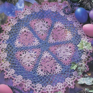 May include: A crochet doily with a delicate floral design in shades of pink, purple, and blue. The doily is surrounded by green leaves and purple flowers. The text "A Touch of Color" and "Tenderness" are visible in the image.