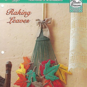 May include: A handmade autumn-themed wall hanging featuring a green plastic canvas broom with a beige bow. Colorful stitched leaves in red, orange, yellow, and green are attached. The text "Raking Leaves" is visible.