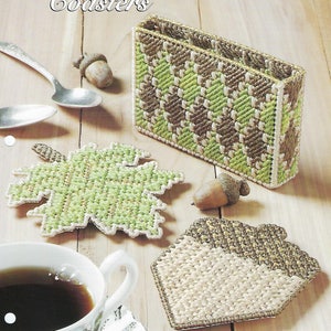 May include: A plastic canvas craft pattern for a set of coasters featuring a green leaf, an acorn, and a brown and green geometric design. The pattern is titled "Golden Accent Coasters" and is part of the "Nature's Wonders" collection.