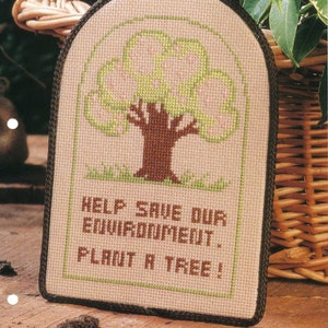 May include: Cross stitch pattern featuring a tree with pink blossoms and the text "Help save our environment. Plant a tree!"  The pattern is framed in a brown border and is surrounded by a wicker basket and green leaves.
