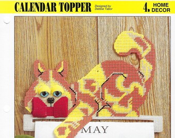 May include: A colorful plastic canvas craft of a cat holding a book with a red bow tie. The cat is holding a calendar with the month of May displayed. The calendar is on a wooden surface.