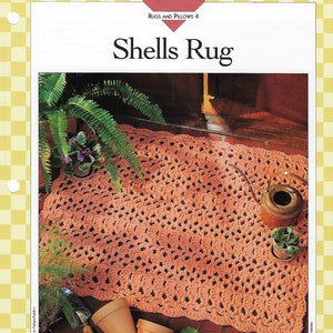 May include: A crocheted rug with a shell stitch pattern in a peach color. The rug is laid out on a tiled floor with potted plants and gardening tools around it. The text "Shells Rug" is at the top of the image.