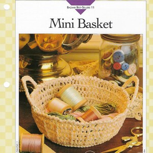 May include: A crocheted mini basket with handles, filled with spools of thread in various colors. The basket is approximately 2 inches deep. The image also includes a jar of buttons and a pair of scissors.