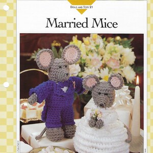 May include: A crocheted wedding couple of mice. The groom is wearing a purple suit and the bride is wearing a white dress. The mice are standing on a table with a bowl of white and green marshmallows.