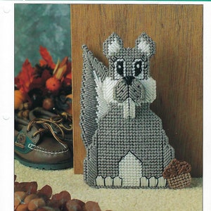May include: A gray and white plastic canvas squirrel doorstop with a brown acorn. The squirrel is sitting on its haunches with its tail curled behind it. The doorstop is designed by Diane Ray.