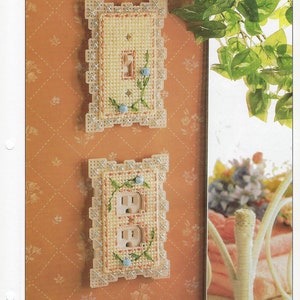 May include: Two plastic canvas wallplate covers with a floral design. The top cover is for a light switch and the bottom cover is for a double outlet. Both covers have a white background with blue and green floral accents.
