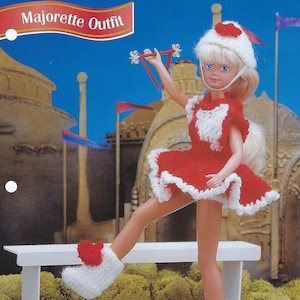 May include: A crocheted doll outfit for a majorette doll. The outfit is red and white with a white hat and white boots. The doll is holding a red and white baton.