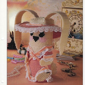 May include: A handmade lop-ear bunny made of plastic canvas, wearing a pink dress with white lace trim and a matching hat. The bunny has black eyes and a pink nose. The design is from Annie's International Plastic Canvas Club.