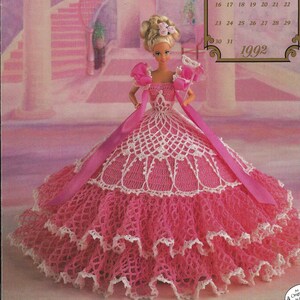 May include: A pink crochet doll dress with white lace trim. The dress is designed for a Barbie doll and features a large, full skirt. The doll is wearing a white headband with pink flowers. The background is a pink and white room with a staircase.