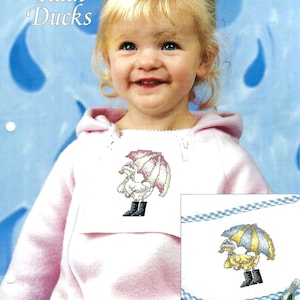 May include: A cross stitch pattern featuring a child wearing a pink sweater with a cross stitch design of a duck wearing a pink and white umbrella. The pattern is titled "Rain Ducks" and is part of the "Cross Stitch Collector's Series" by Butterfly Kisses.
