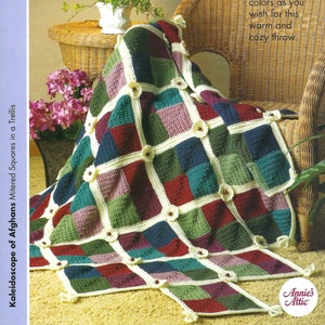 May include: A crochet blanket with a trellis pattern in shades of green, red, blue, and purple. The blanket is made of squares with white crocheted flowers in the center of each square. The blanket is draped over a wicker chair with a plant in a white pot on a wicker table.