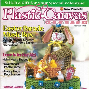 May include: A magazine cover for "Plastic Canvas Crafts" featuring an Easter-themed music box. The music box is a bunny with a basket, chicks, and decorative eggs. The cover text reads "Easter Parade Music Box" and "February 1998."