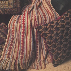 May include: A crocheted throw blanket with a striped pattern in shades of beige, red, and blue. The blanket is draped over a couch with two pillows featuring geometric patterns in shades of brown, beige, and red. The text "ARROWHEAD AFGHAN" is visible in the top center of the image.