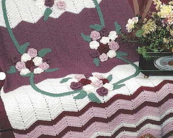May include: A crocheted floral ripple afghan in shades of purple, pink, and white. The afghan features a repeating chevron pattern and a floral design with roses and leaves.