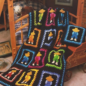 May include: A black crochet blanket with colorful squares featuring a cartoon figure wearing overalls and a hat. The blanket is on a wooden floor with a wooden toy wagon and a stuffed animal.