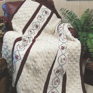 May include: A white crocheted throw blanket with a brown border and floral embroidery. The blanket is draped over a brown leather armchair with tassels hanging from the bottom edge. The text "Swiss Holiday Afghan" is printed above the image.