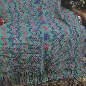 May include: A crocheted throw blanket with a raspberry ripple pattern in shades of green, pink, and purple. The blanket has a fringe border and is draped over a chair.