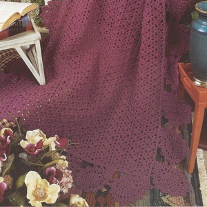 May include: A crocheted throw blanket in a deep burgundy color with a lacy pattern. The blanket is draped over a white shelf and a wooden table with a book on top. The text "Pineapples & Lace" is visible in the top right corner of the image.