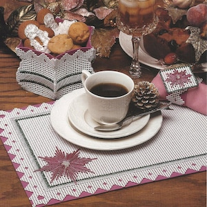 May include: A white plastic canvas craft project featuring a poinsettia design. The project is a placemat with a pink border and a green and white poinsettia in the center. The image also shows a cup of coffee, a glass of iced tea, and a basket of cookies.