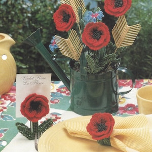 May include: A plastic canvas craft pattern for a wheat field harvest. The pattern features a bouquet of red poppies, wheat stalks, and blue flowers in a green watering can. The pattern also includes instructions for making a poppy flower napkin ring and a poppy flower place card holder.
