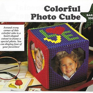May include: A colorful photo cube with heart-shaped cutouts on each side. The cube is made of plastic canvas and is decorated with flowers and hearts. The cube is a craft project and is perfect for displaying photos of loved ones.
