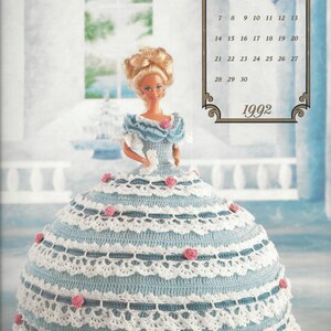 May include: A crocheted doll wearing a blue and white dress with a lace trim. The doll has blonde hair and blue eyes. The doll is standing in front of a window with a calendar that says "June 1992".