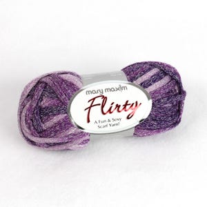 May include: A skein of Mary Maxim Flirty scarf yarn in shades of purple and silver. The yarn is labeled "Flirty" with the tagline "A Fun & Sexy Scarf Yarn!" The yarn is a blend of cotton and dye lot.