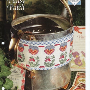 May include: A cross stitch pattern for a pansy border, featuring a blue and white gingham background with a row of pink and purple pansies. The pattern is titled "Pansy Patch" and is part of the "Heart Songs Cross Stitch Collector's Series".