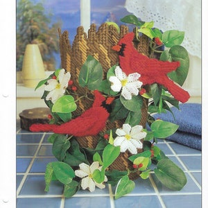 May include: A plastic canvas craft project featuring two red cardinals perched on a woven brown basket with white flowers and green leaves. The project is titled "Cardinals Renuzit Cover 4. Home Decor".