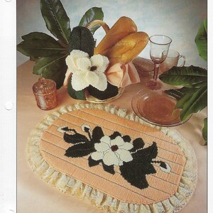 May include: A plastic canvas table runner with a peach background and a white magnolia flower with green leaves. The runner has a lace trim and is designed for home decor.
