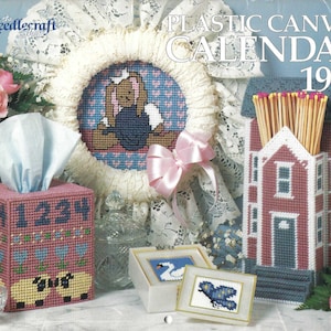 May include: Plastic canvas craft patterns for a calendar, tissue box cover, and match holder. The calendar is a house with a red roof and blue trim. The tissue box cover has a pink base with a sheep design. The match holder is a house with a red roof and blue trim. The patterns are from 1990.