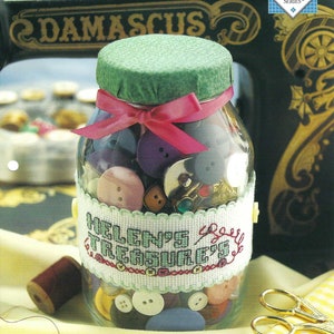 May include: A glass jar filled with colorful buttons, with a green lid and a pink ribbon. The jar has a white cross-stitch design around it that says "Helen's Treasures". The jar is sitting on a yellow and white striped fabric.