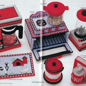 May include: A collection of handmade plastic canvas kitchen toys, including a coffee maker, stove, blender, soup pot, popcorn popper, place mat, and electric can opener. The toys are primarily red, white, and black, with some gray and blue accents. The place mat features a farm scene.