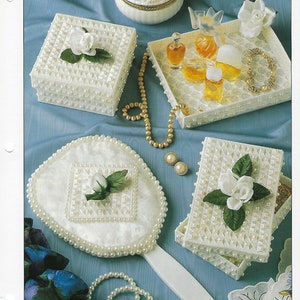 May include: A white plastic canvas vanity set with pearl accents. The set includes a tray, two boxes, and a hand mirror. The boxes and tray feature a white rose design. The mirror has a heart-shaped frame and a white rose design in the center.