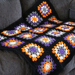 May include: A handmade crochet blanket in a granny square pattern. The blanket features a black background with squares of white, gray, orange, and purple. The blanket is draped over a gray couch.