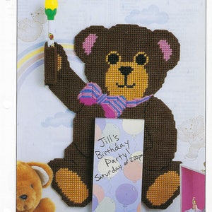 May include: A brown teddy bear with a pink bow tie holds a white marker with a yellow flower on top. The bear is holding a white card with the text "Jill's Birthday Party Saturday at 2:00pm" written on it. The card is decorated with colorful balloons.