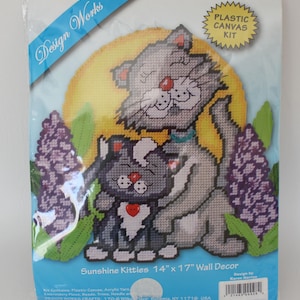 May include: A plastic canvas kit for creating a wall decor featuring two grey and white kittens with a red heart, surrounded by purple flowers and a yellow sun. The kit includes acrylic yarn, embroidery floss, beads, trims, a needle, and plastic canvas. The finished piece measures 14 inches by 17 inches. The kit is from Design Works Crafts.