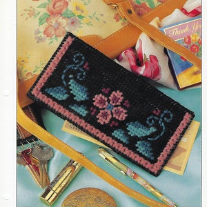 May include: A black plastic canvas checkbook cover with a pink and blue floral design. The cover is lying on a light blue surface with a brown leather purse, a gold lipstick tube, and a gold pen.