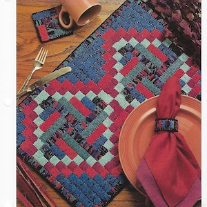 May include: A set of four placemats with a log cabin design. The placemats are made of plastic canvas and are woven with red, blue, green, and white yarn. The placemats are shown on a wooden table with a red plate, a red napkin, and a brown mug.