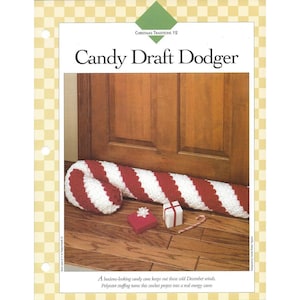 May include: A crochet candy cane draft dodger with red and white diagonal stripes. The text reads "Christmas Traditions 12" and "Candy Draft Dodger". Two small gift boxes and a miniature candy cane are placed in front of the door draft stopper.