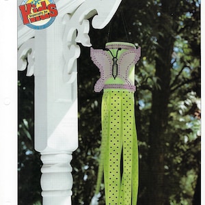 May include: A green and purple butterfly windsock with white polka dots hanging from a white porch post. The windsock is made of plastic canvas and has a butterfly design with black accents. The windsock is labeled "Quick & Easy Creations" and "Especially for Kids".