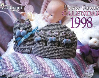 May include: A gray plastic canvas calendar cover with cat designs, blue pom-poms, and a blue ribbon. The calendar is dated 1998. The cover is on a pillow with a striped pattern in shades of blue, purple, and white.