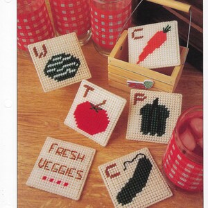 May include: Set of six white plastic canvas coasters with cross-stitch designs of vegetables. The coasters feature a watermelon, tomato, carrot, pepper, cucumber, and a sign that says "Fresh Veggies".