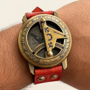 May include: A brass sundial watch with a red leather strap featuring two metal rivets. The circular face displays Roman numerals, a central gnomon with a sun engraving, and the text Ross London. A compass is visible beneath the gnomon.