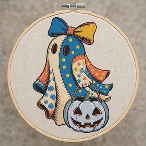 Op de afbeelding: Een borduurring met een stoffen paneel met een kleurrijke spookillustratie. Het spook draagt een grote blauwe en gele strik en draagt een lichtblauwe pompoenemmer. Het spook heeft blauwe stippen en bloemmotieven.